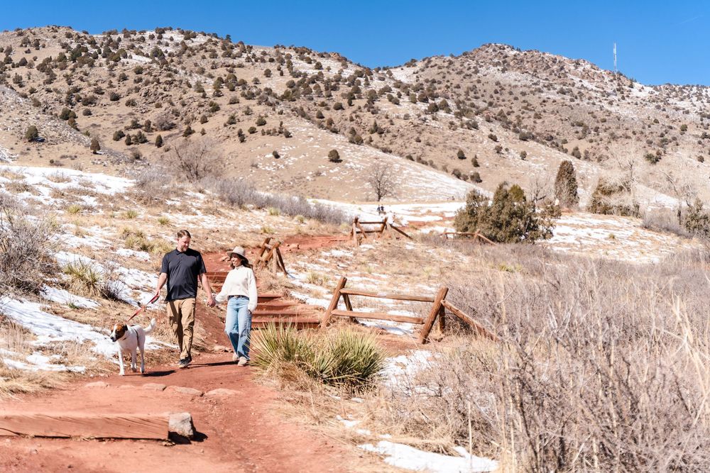 Couple walking dog on red dirt trail through snowy mountain landscape with scrub brush and hills in background.