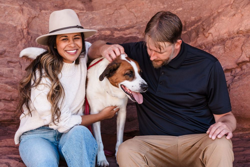 A smiling couple sits against red rock with a happy Jack Russell Terrier dog between them outdoors.