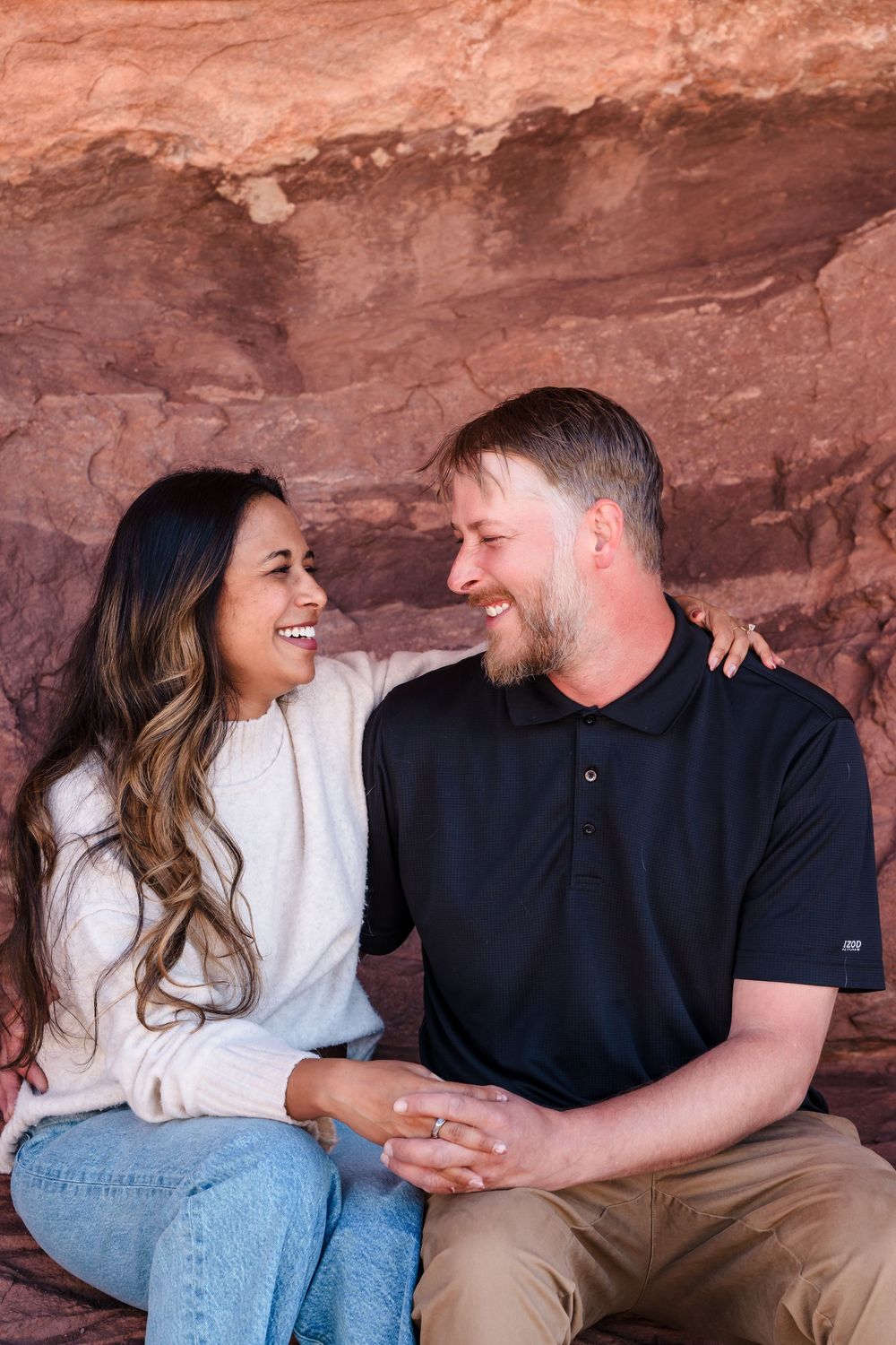 Engaged couple sitting and laughing together against a red sandstone rock wall at Red Rocks Park.