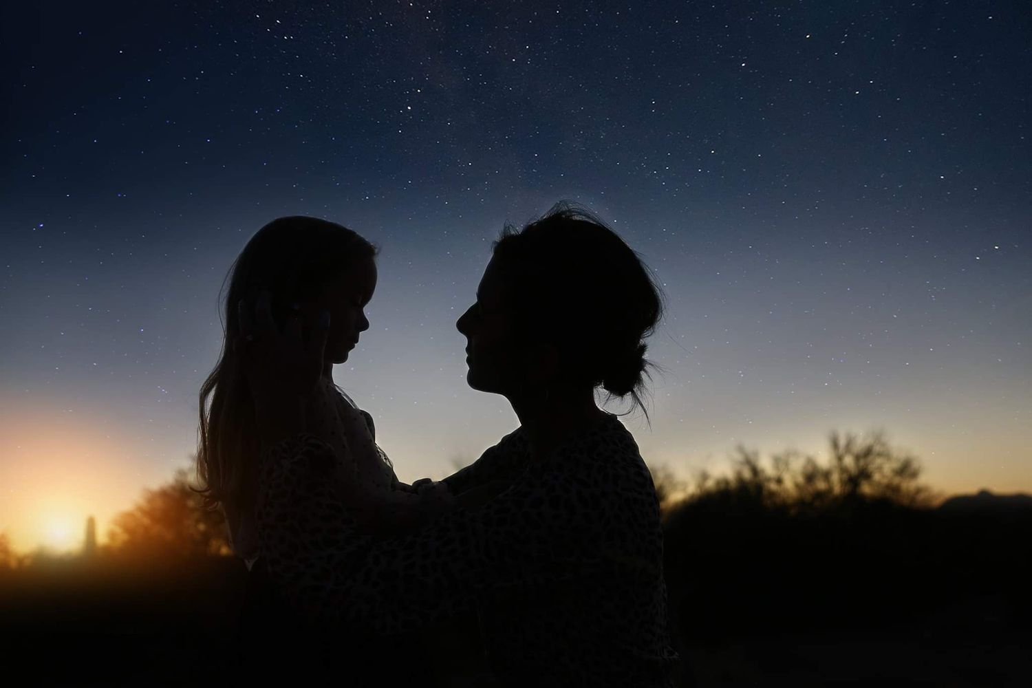 Silhouette of a mother and child gazing at each other under a starry twilight sky with a warm glow on the horizon.