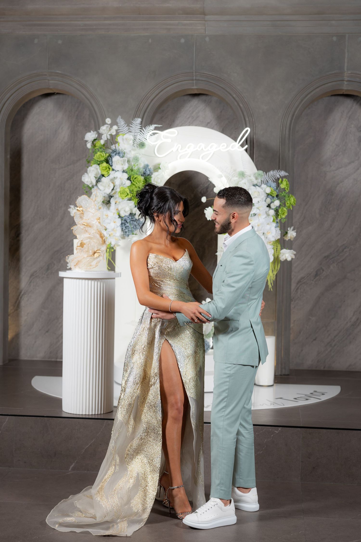 Bride and groom holding hands before floral arch with neon sign in elegant venue.