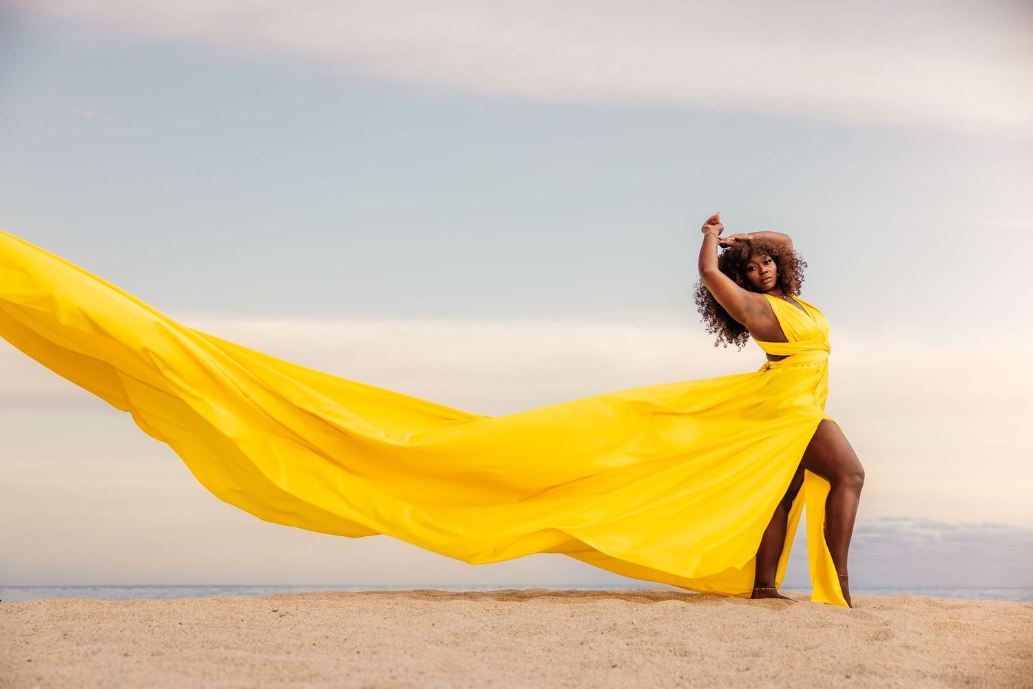A Black woman in a flowing yellow dress poses on a sandy beach, her fabric billowing dramatically in the wind.