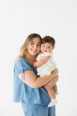 Smiling young mother in blue dress holding happy baby girl in white outfit against white background.