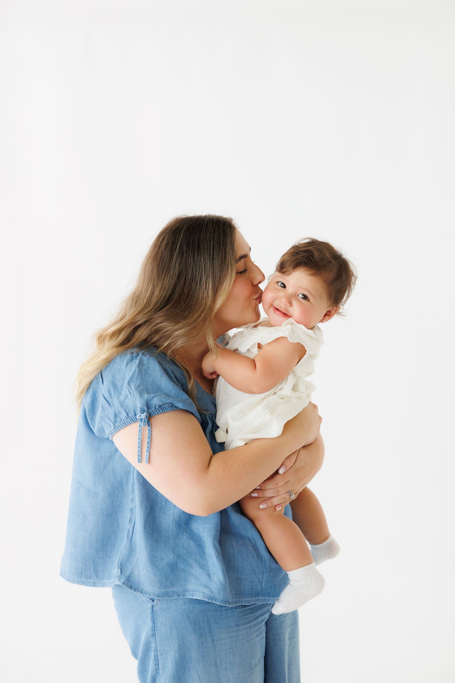 Mother in blue dress kissing her smiling baby girl in white outfit against white background.
