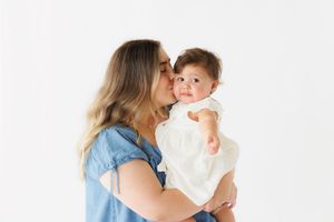 A mother in a denim shirt kisses her baby girl in a white dress against a clean white background.