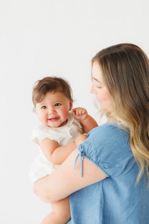 A smiling baby in a white dress being held by a woman in a blue denim outfit against a white background.