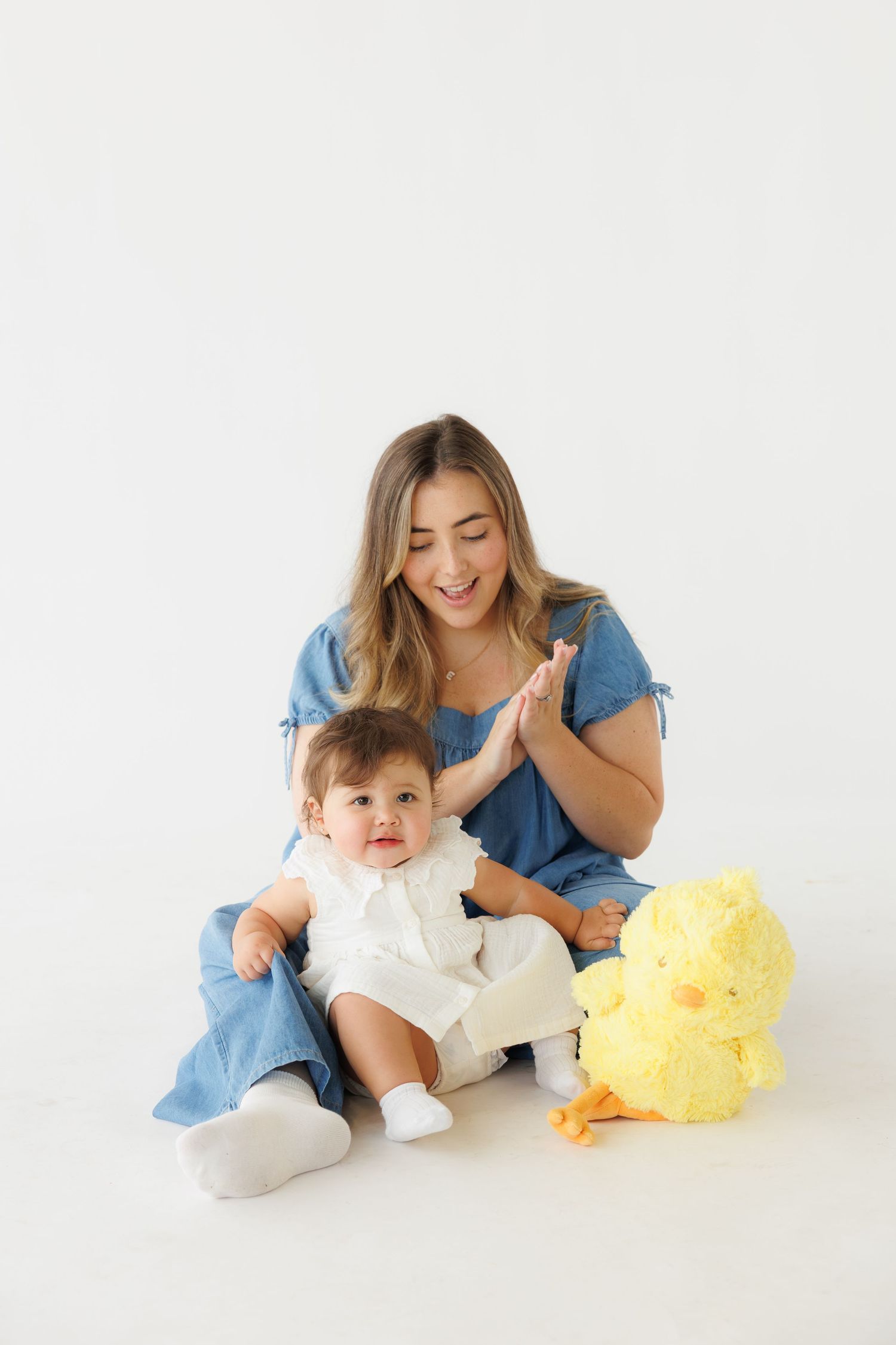 A smiling mother in blue dress clapping hands with her baby in white outfit, seated next to a yellow duck plush toy.