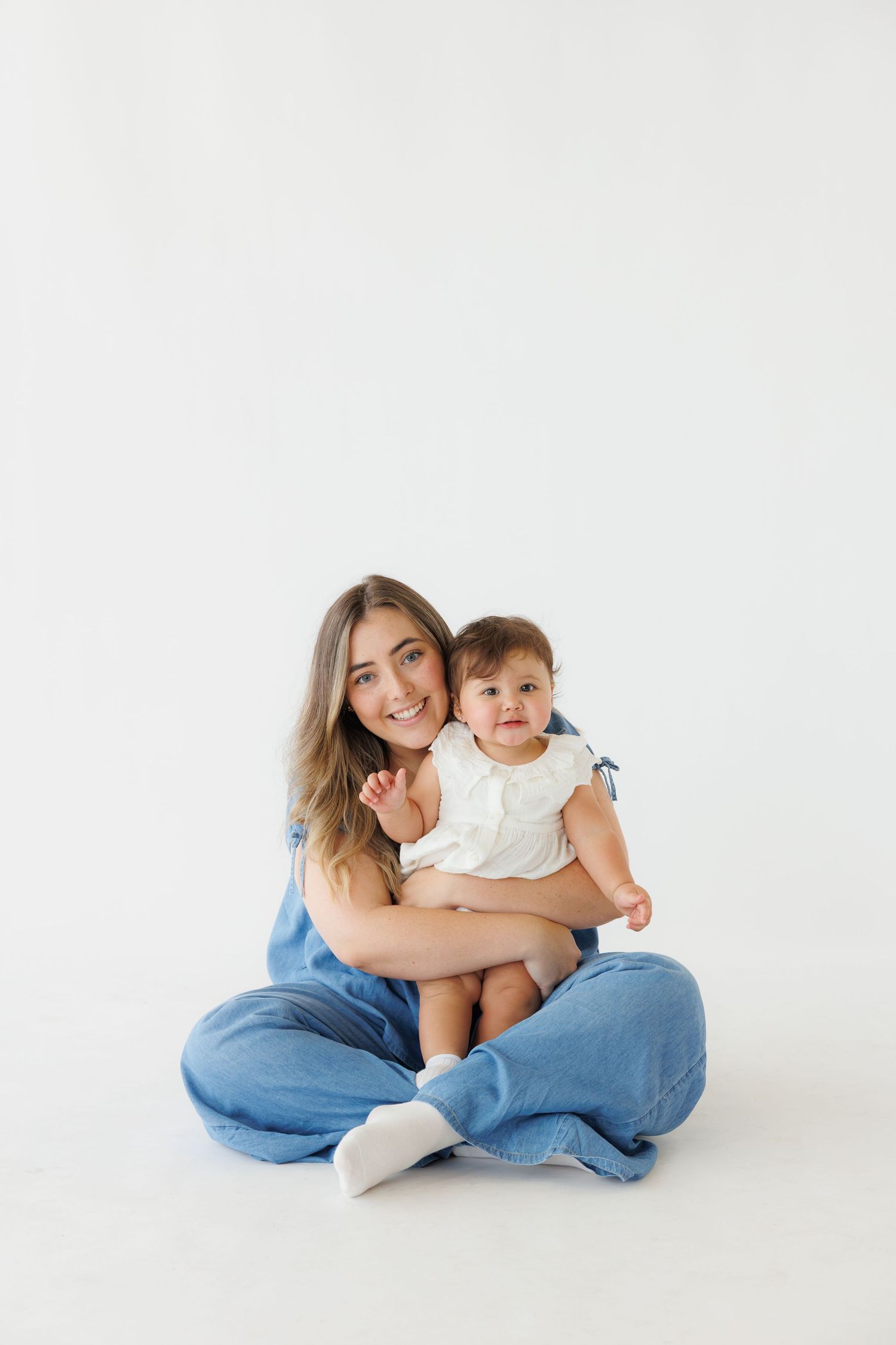 Smiling mother in blue jeans sitting cross-legged holding baby girl in white dress on white background.