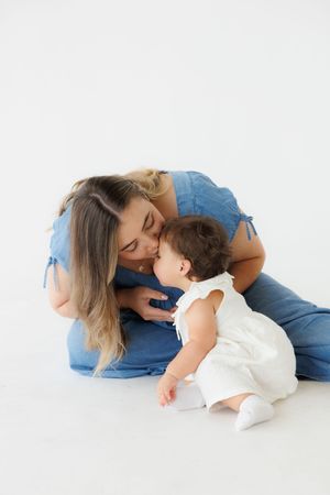 A mother in blue denim gently kisses her baby girl in a white dress while sitting on a white studio floor.