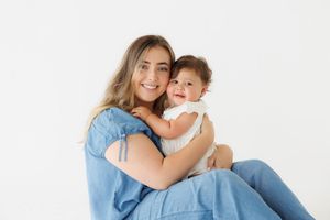 Young woman in blue denim outfit sitting and cuddling smiling baby in white romper on white background.