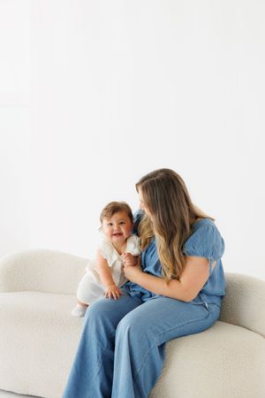 A mother in a blue jumpsuit sitting on a white sofa, lovingly holding her smiling baby in a white outfit.