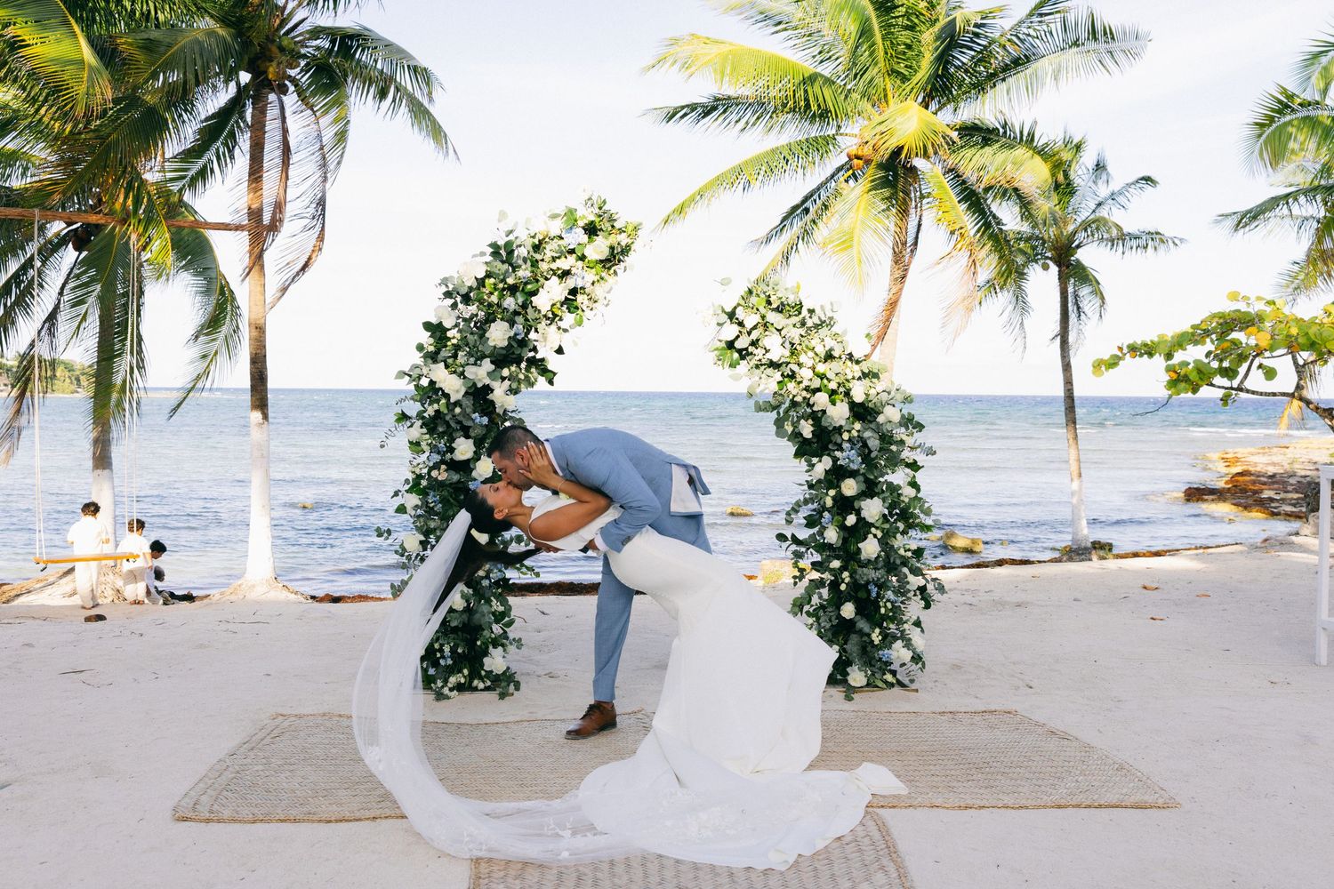 Couple shares romantic kiss under floral arch on tropical beach wedding ceremony with palm trees and ocean backdrop.