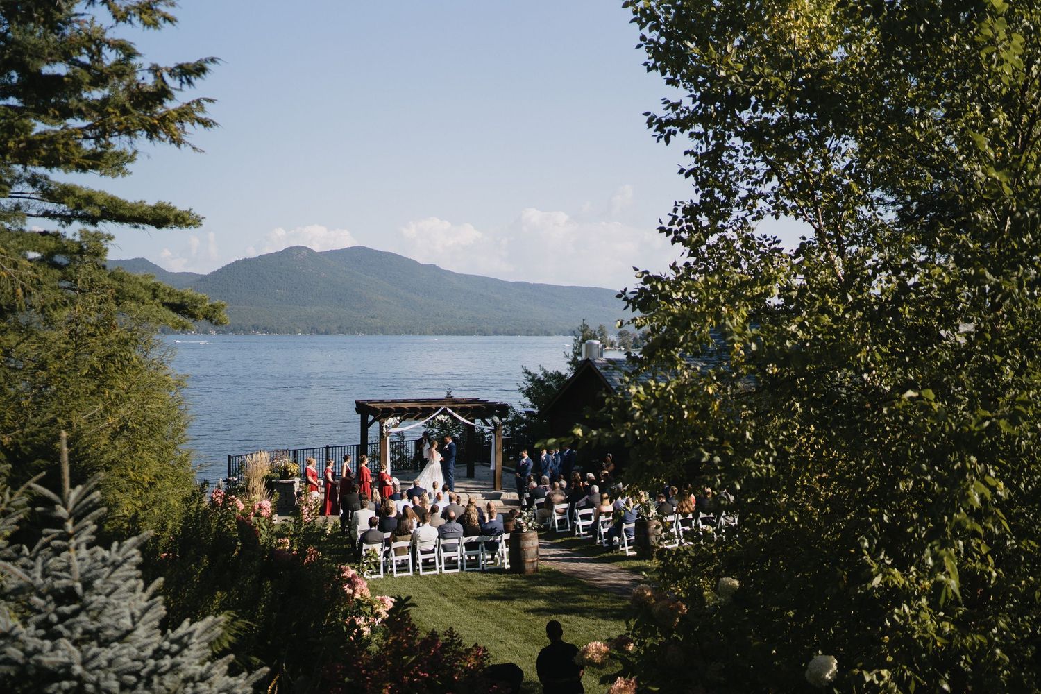 Outdoor lakeside wedding ceremony with mountain views, guests seated on white chairs, surrounded by lush trees.
