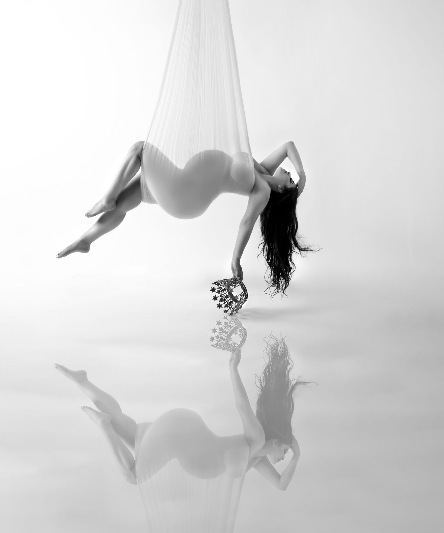 Pregnant woman posing gracefully in aerial silks in a black and white maternity photo.