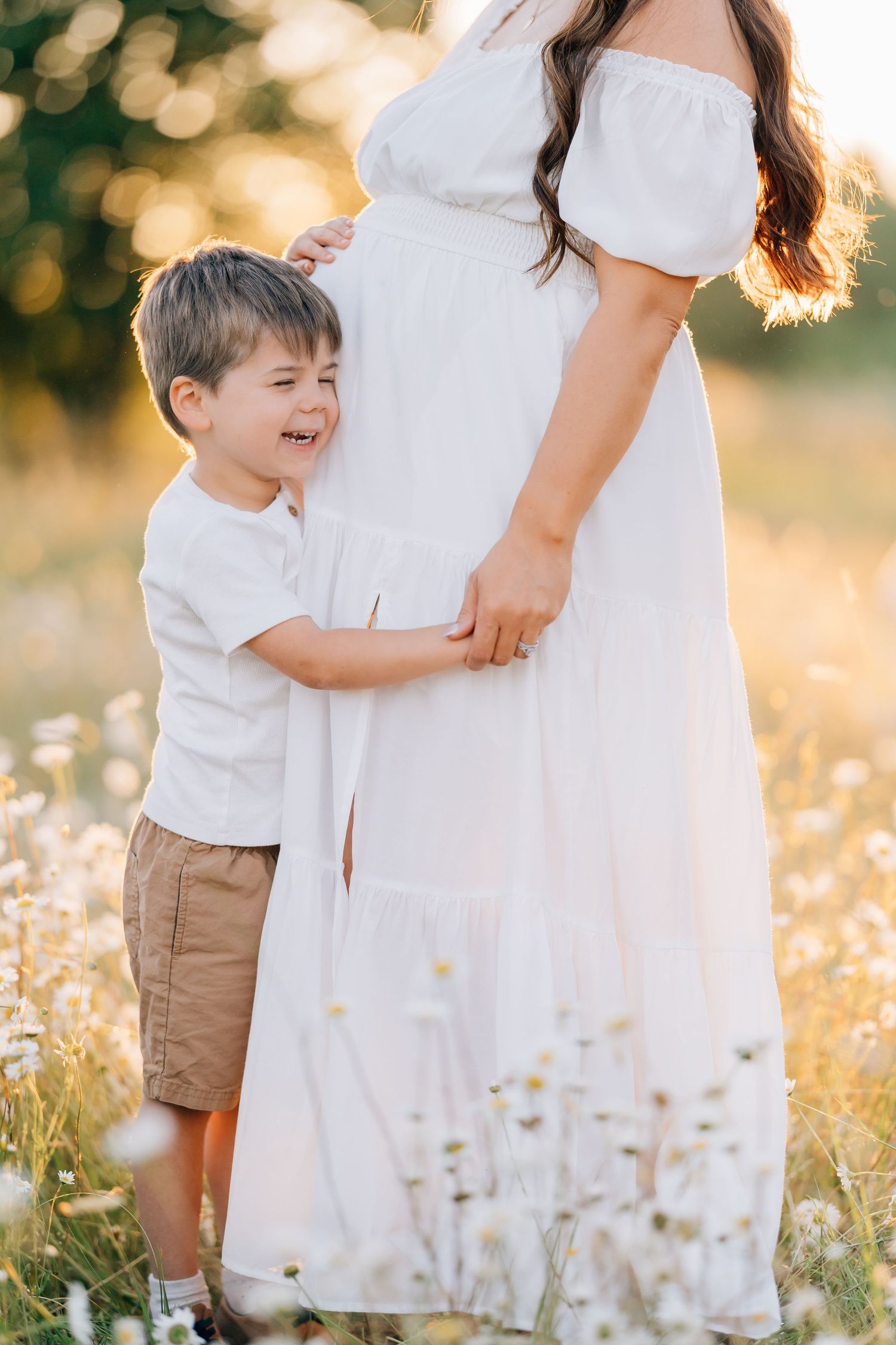 A young boy hugs his pregnant mother in a white dress standing in a sunny daisy field.