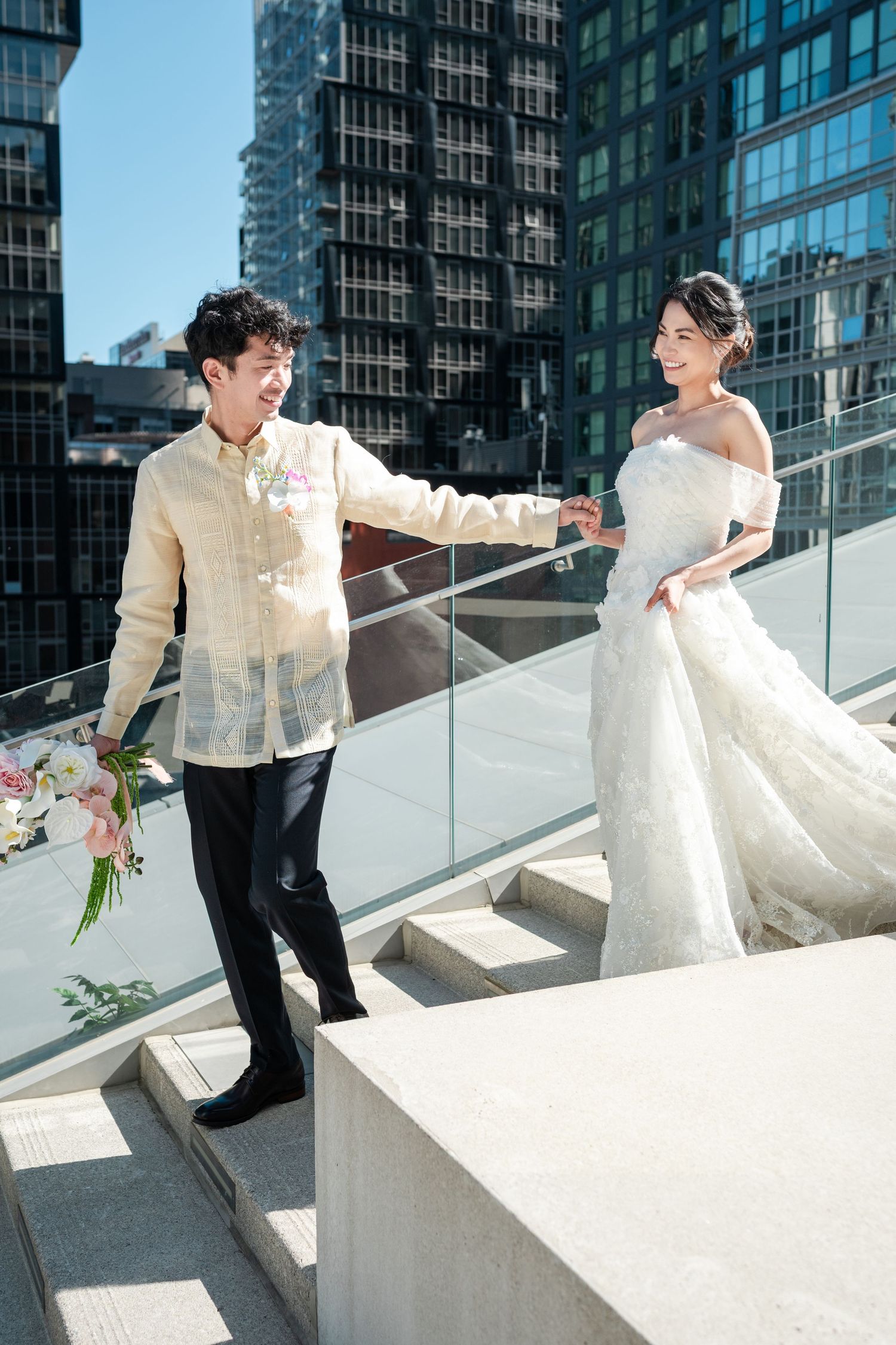 Bride and groom holding hands walking up outdoor stairs with modern city buildings in the background on their wedding day.