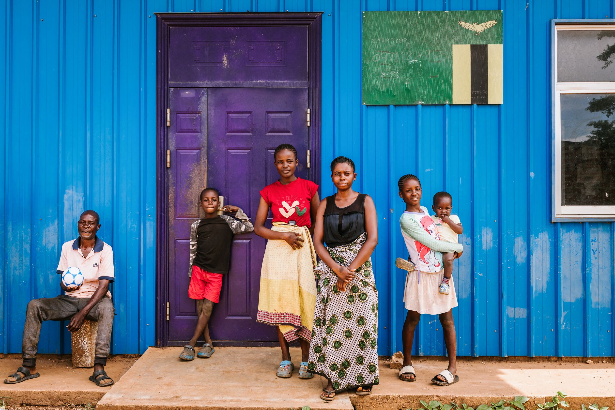 A family of six poses outside a bright blue building with a purple door in rural Africa.