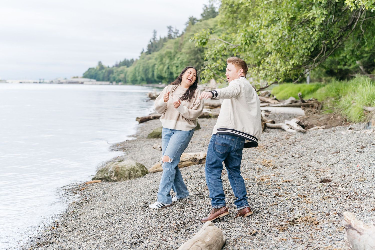 Couple dancing and laughing on a rocky Pacific Northwest beach shoreline surrounded by lush green trees.