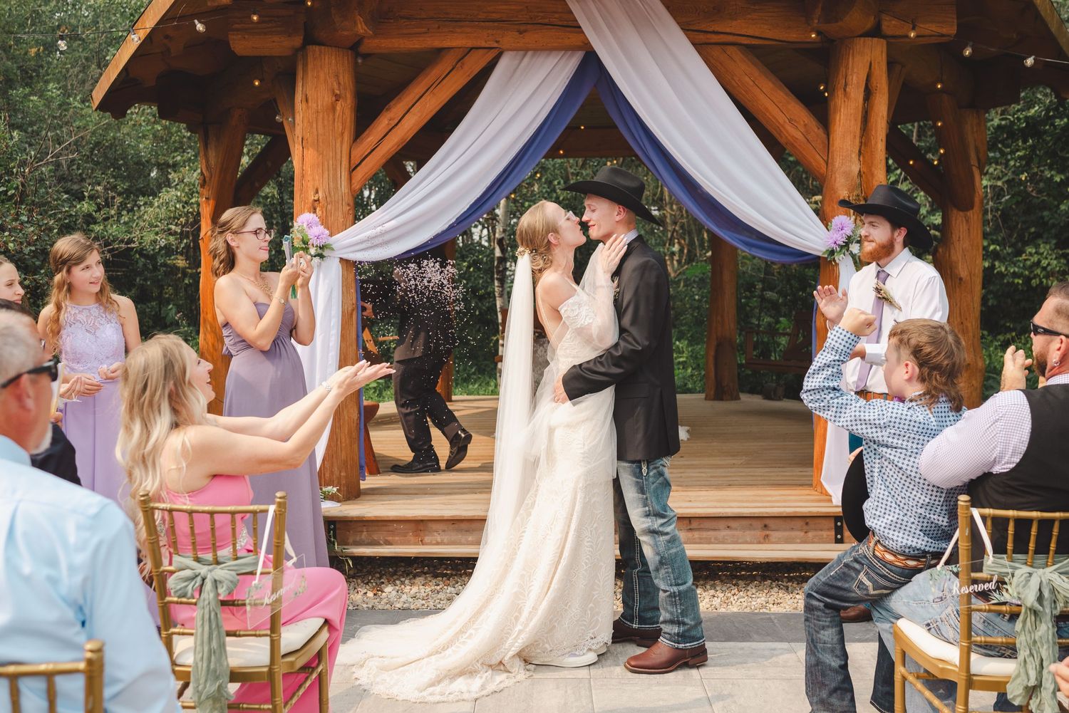 Outdoor wedding ceremony with couple sharing first kiss under a floral arch, surrounded by guests in a rustic garden setting.