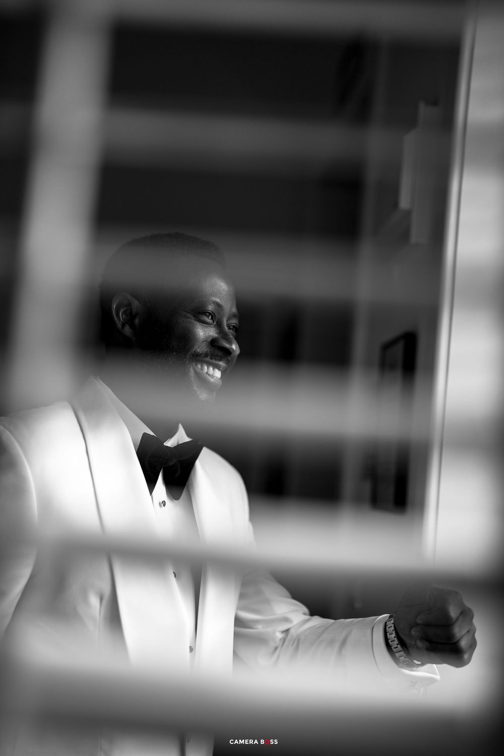 Black and white photo of a smiling groom in a white tuxedo and bow tie, captured through a window frame.