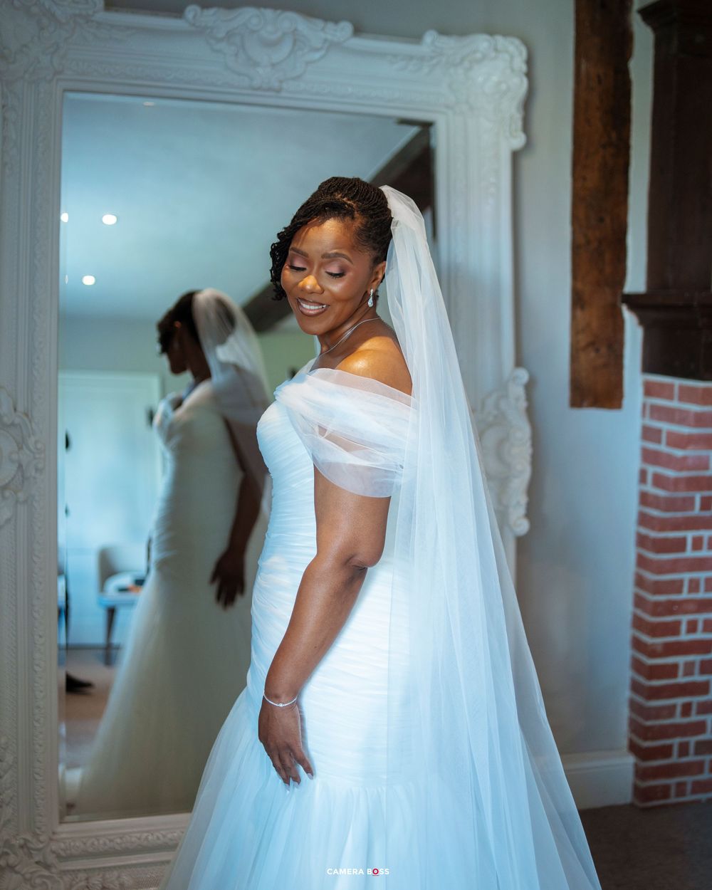 Smiling bride in white off-shoulder wedding gown and veil poses in front of ornate mirror in elegant room.