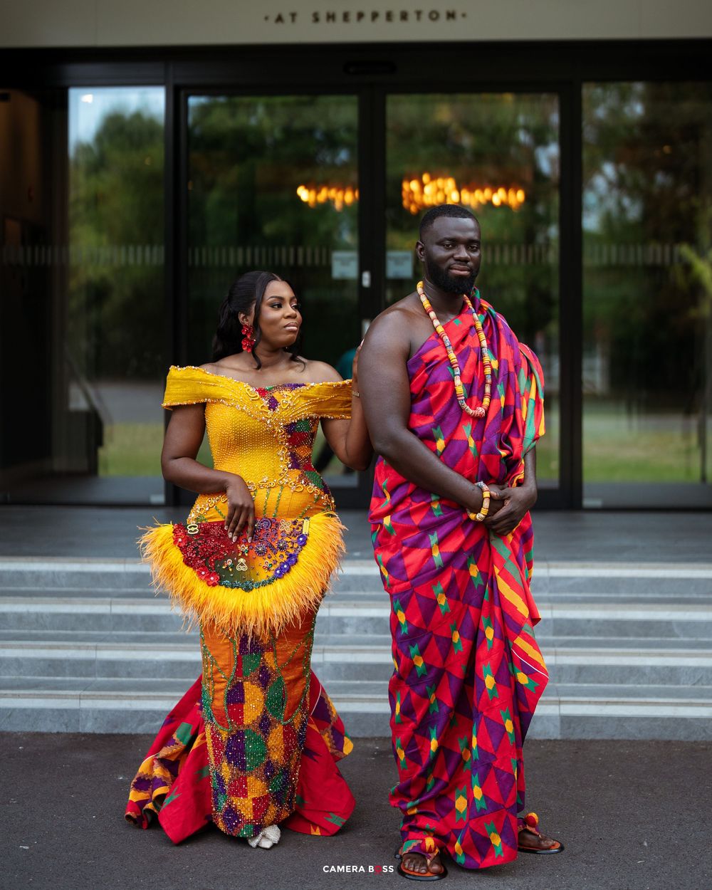 Couple in vibrant traditional African Kente attire posing at Shepperton venue, woman in yellow gown, man in colorful wrap.