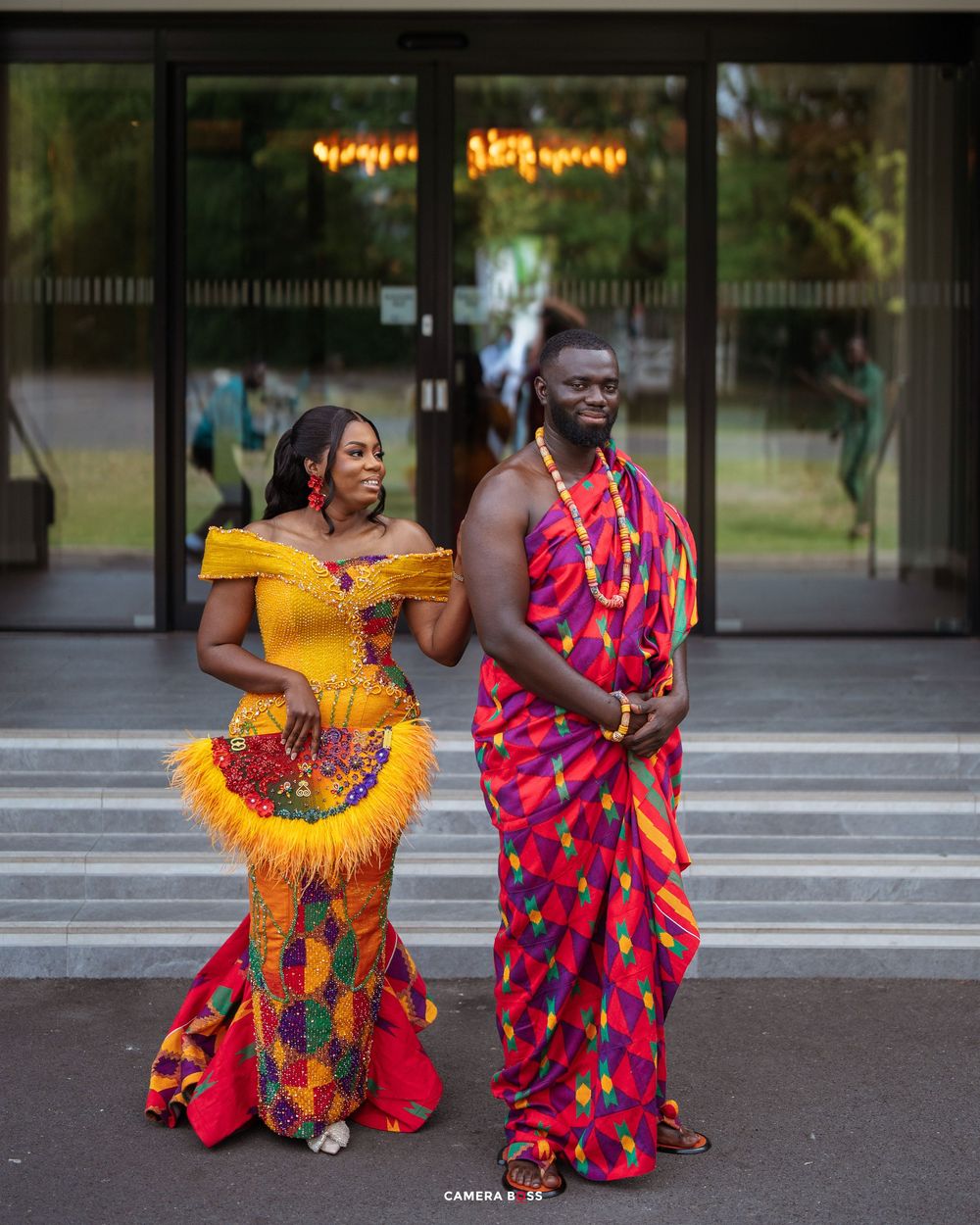 Couple in vibrant traditional African Kente attire posing elegantly at a cultural ceremony outdoors.