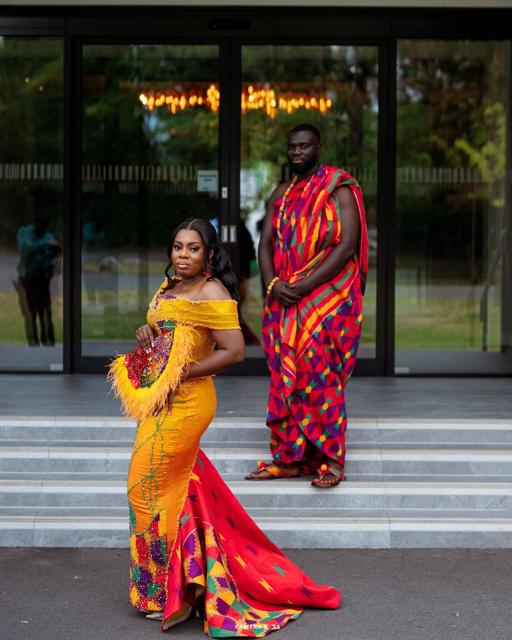 Couple in vibrant African Kente cloth attire posing on steps, woman in yellow gown, man in red patterned wrap.