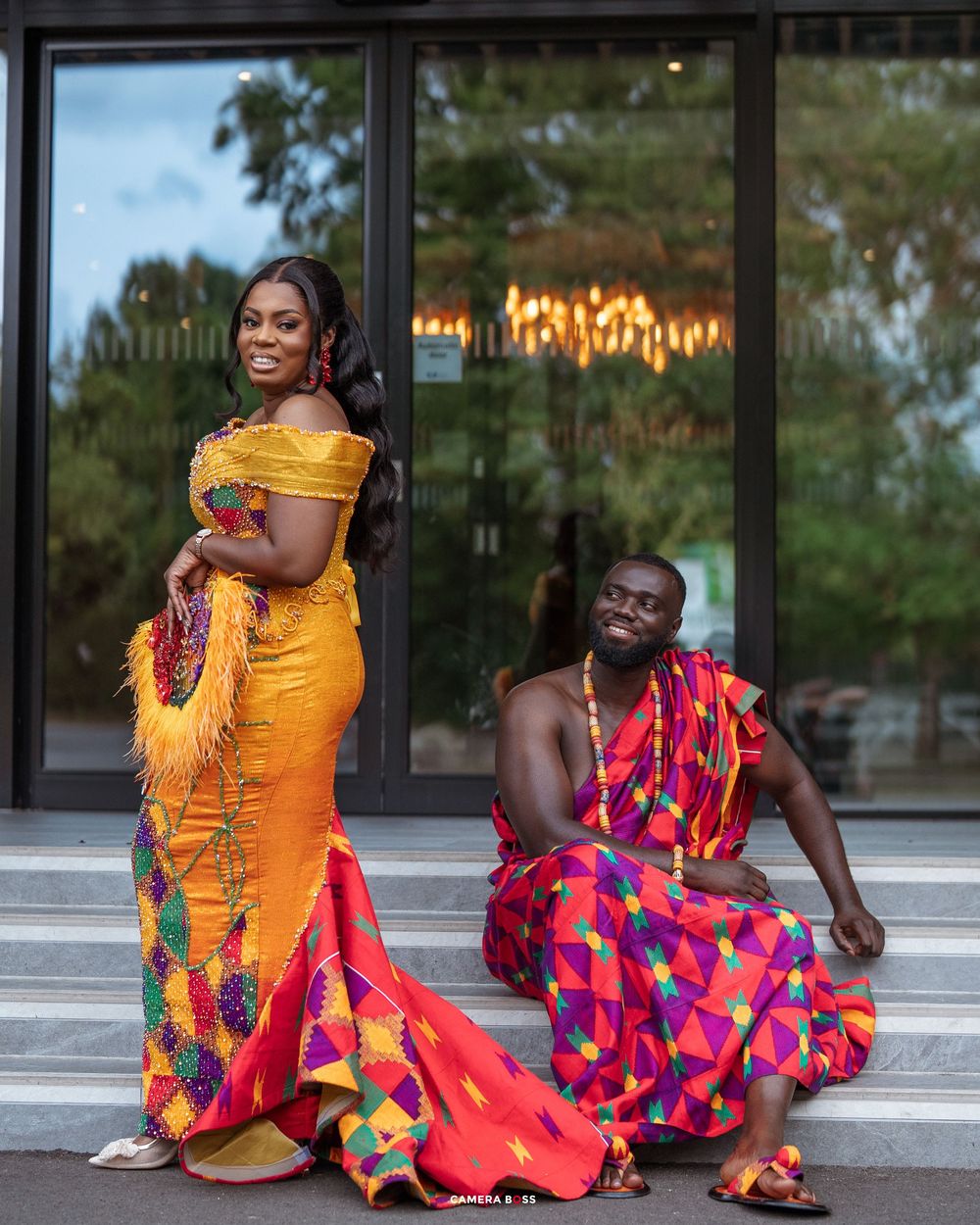 Couple in vibrant Kente cloth traditional Ghanaian attire, woman in yellow mermaid gown, man seated in colorful fabric.