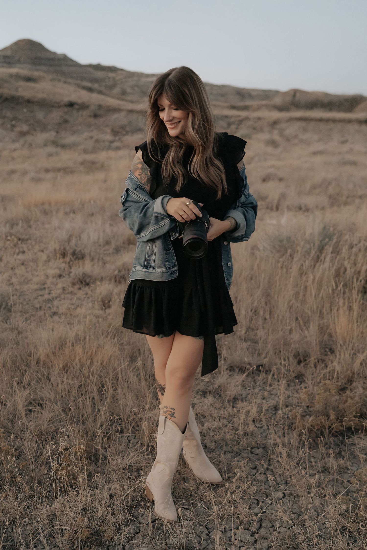 Woman in black dress, denim jacket, and cowboy boots holding a camera in a dry grassland landscape.