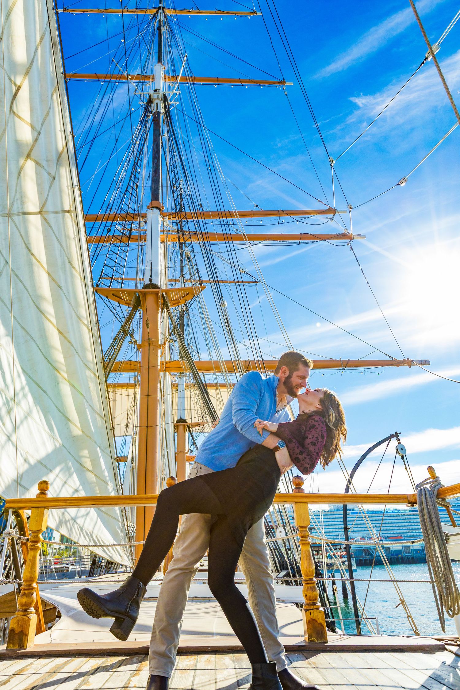 Couple sharing a romantic dip kiss on the deck of a tall sailing ship with masts and blue sky backdrop.