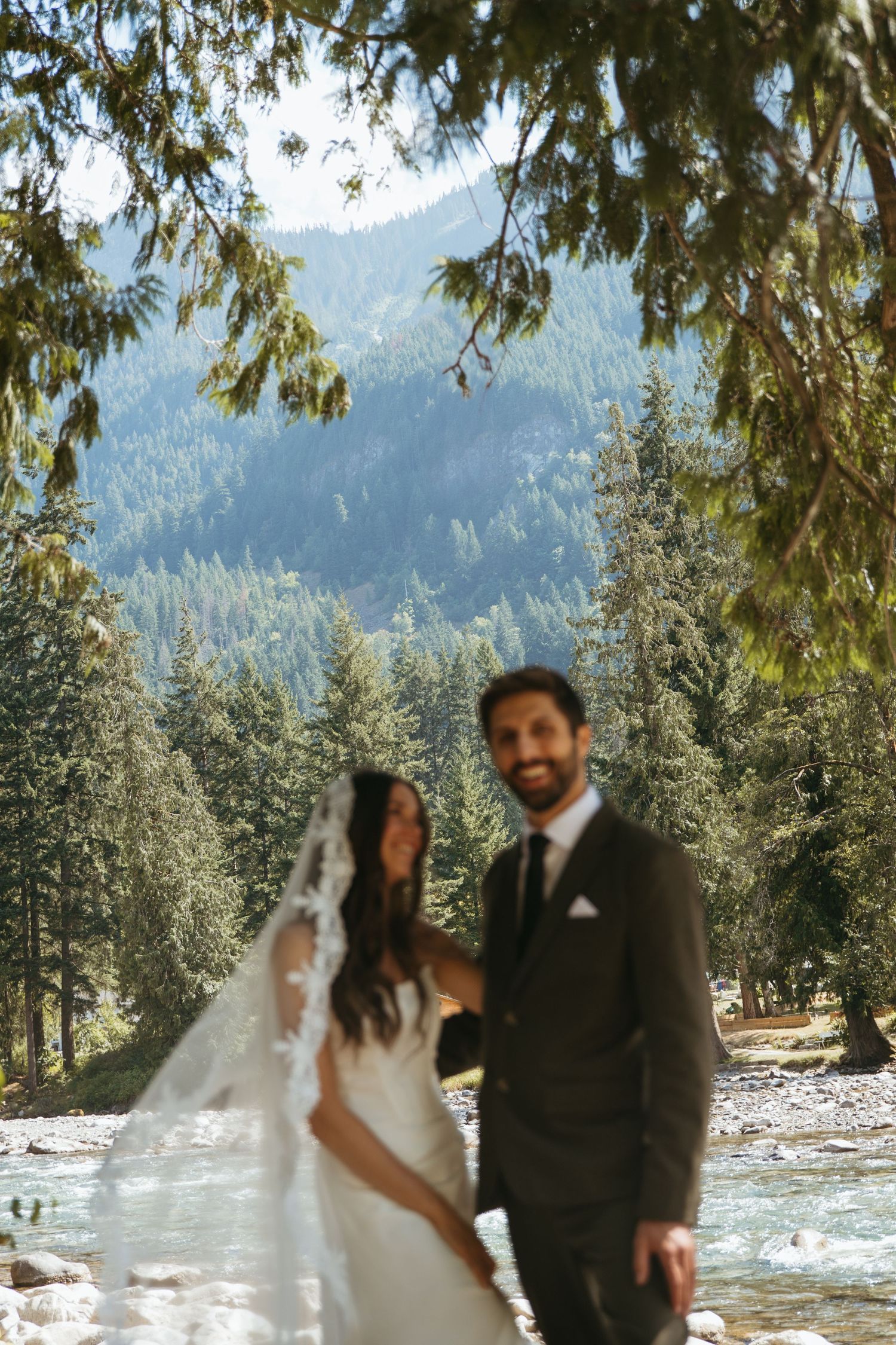 Bride and groom standing by mountain river surrounded by evergreen forest.