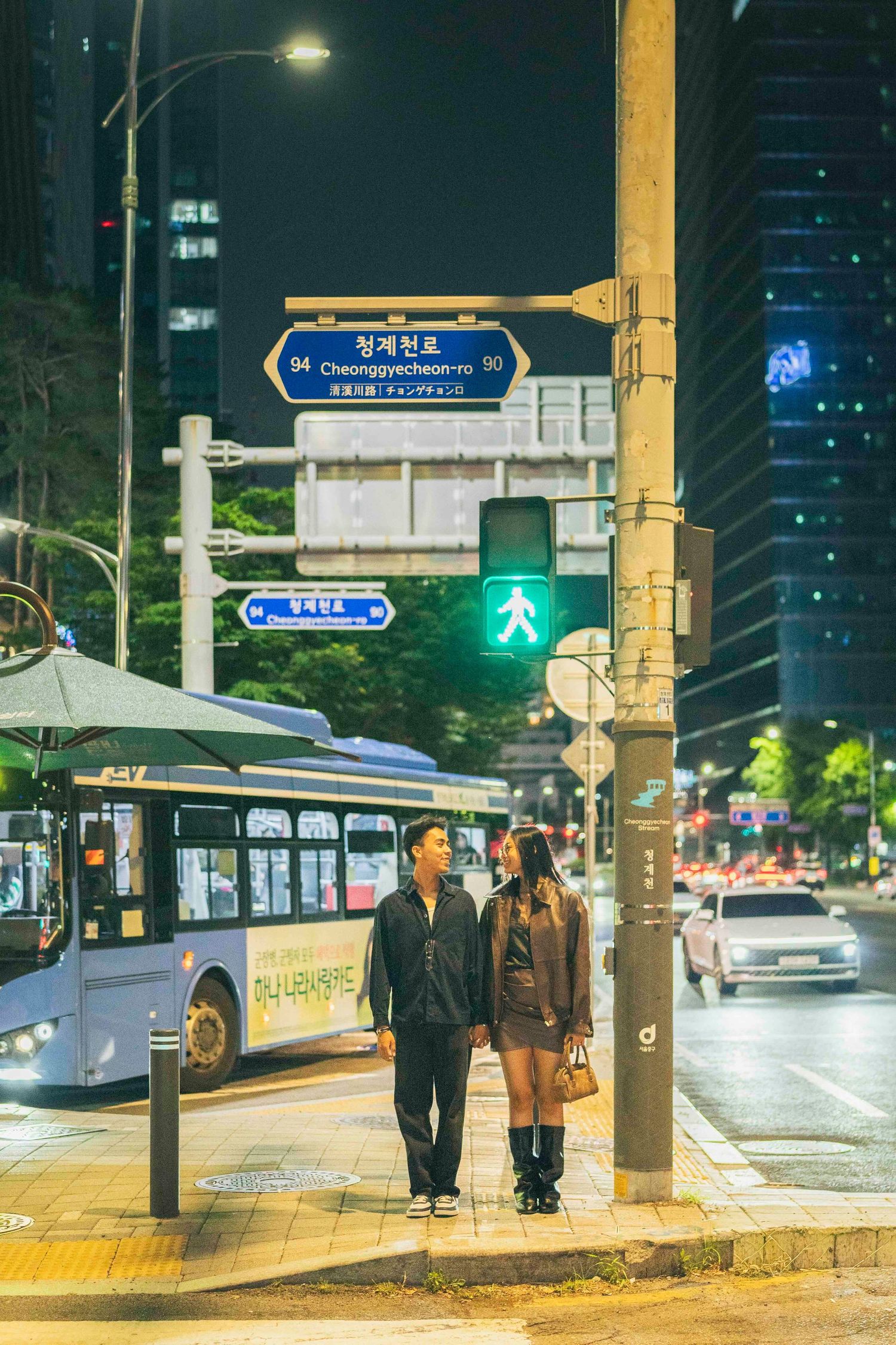 Couple standing at a crosswalk at night in Seoul, South Korea, with city lights and a bus in the background.