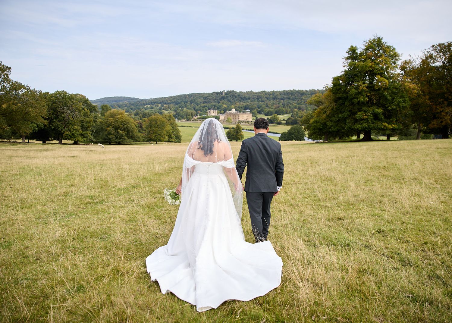 Bride and groom walking hand in hand through a scenic green meadow with rolling hills and trees in the background.