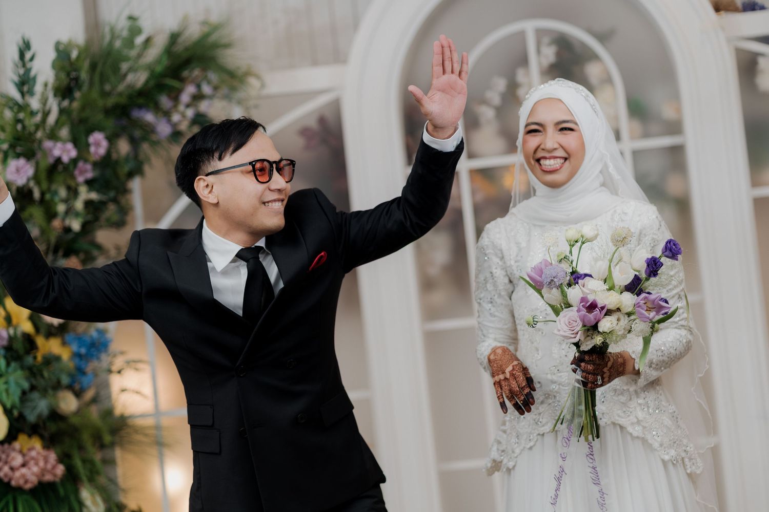 Bride in white hijab holding bouquet stands with groom in black suit waving at wedding ceremony.