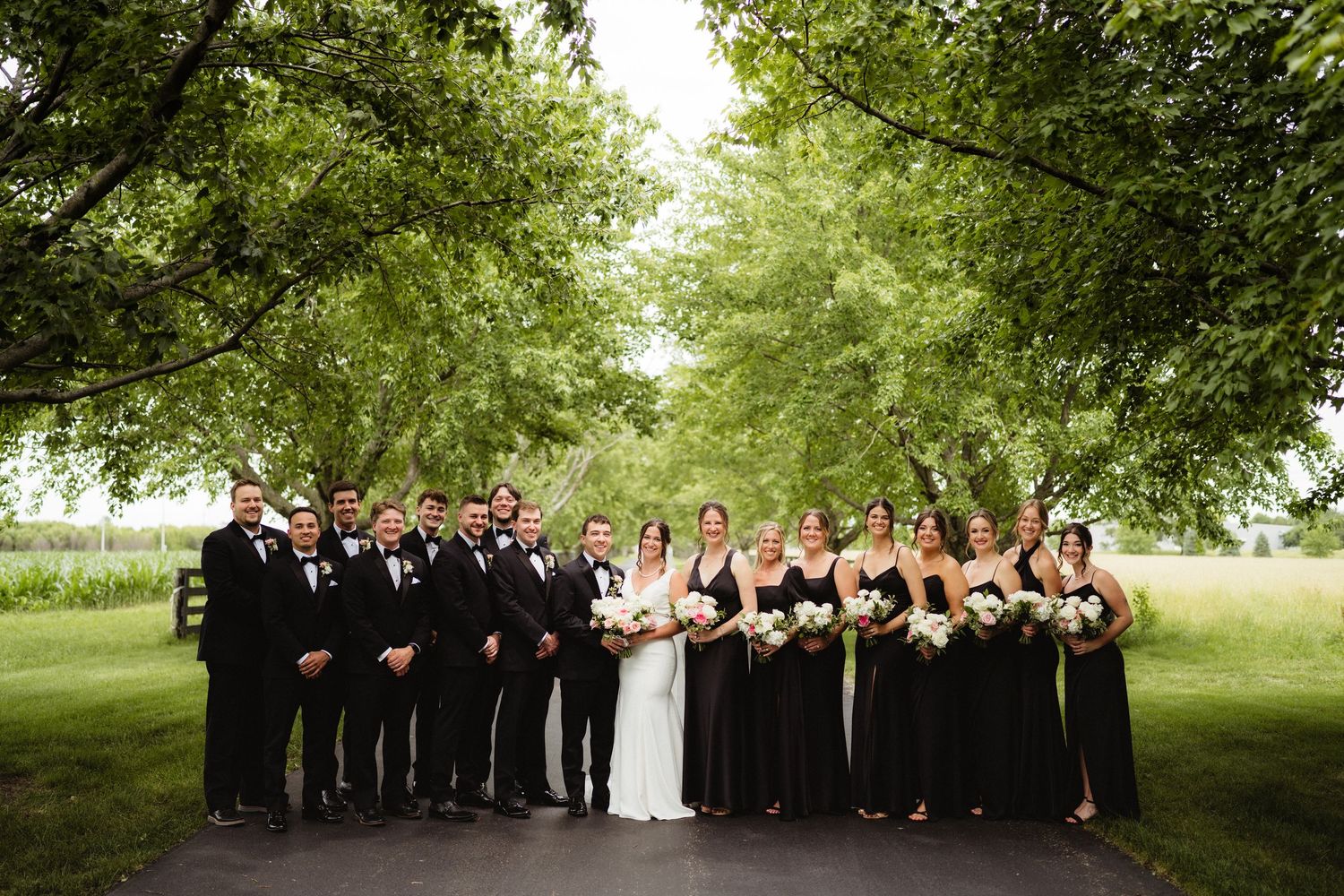 Wedding party in black attire poses outdoors under lush green trees on a paved path.