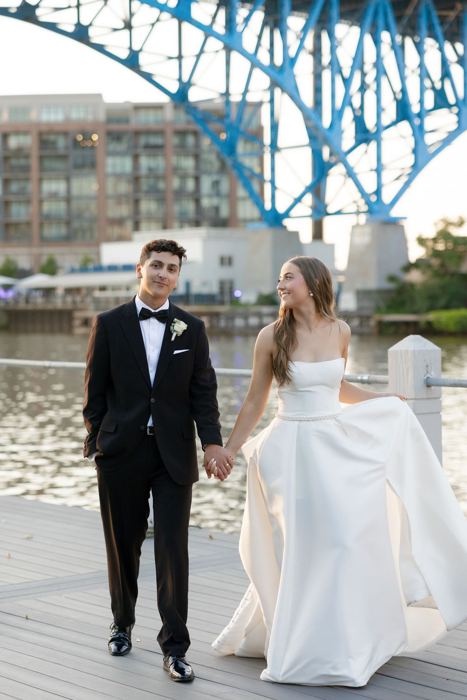 Bride and groom walk hand in hand on waterfront dock beneath a blue bridge.