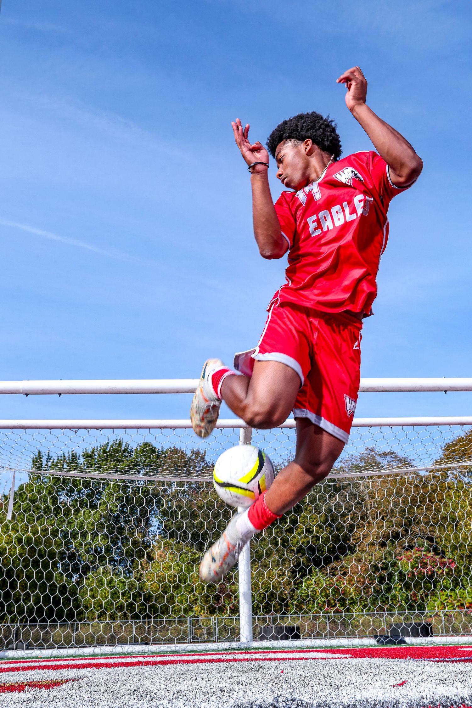 A soccer player in a red Eagles uniform jumps mid-air to kick a ball in front of a goal net under a blue sky.