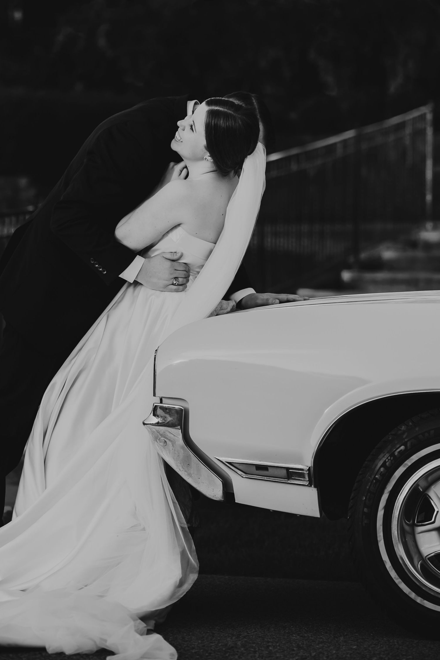 Black and white photo of a bride in a white gown embracing her groom beside a classic white convertible car.