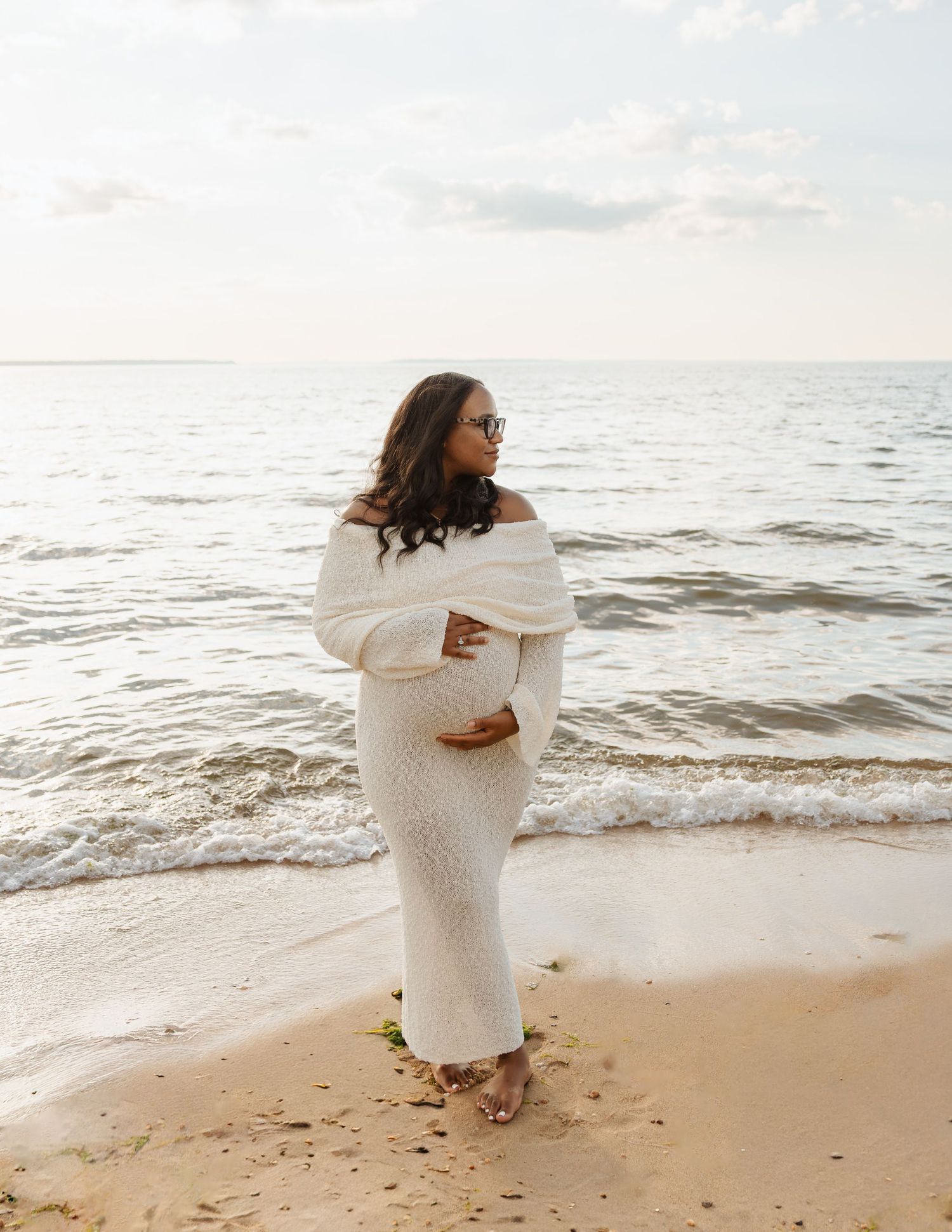 Pregnant woman in white off-shoulder knit dress standing barefoot on sandy beach, gazing at calm ocean waves.