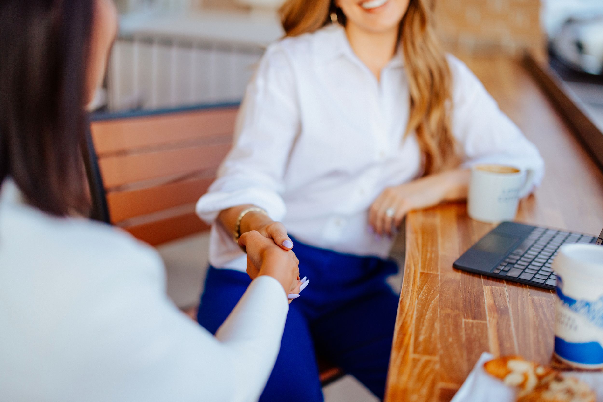Two professional women in white blouses shaking hands over a wooden table with a laptop and coffee cup.