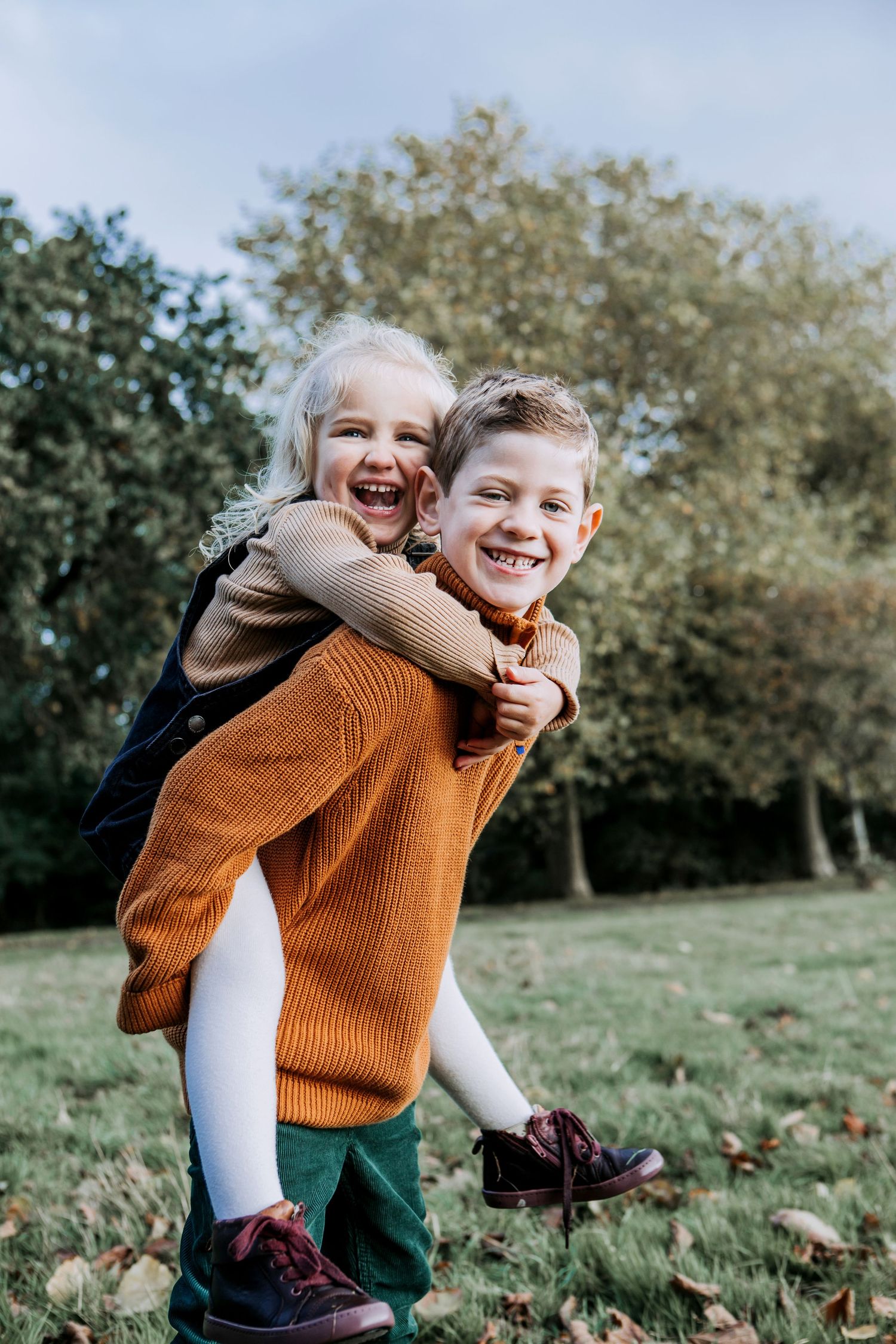 Laughing brother giving his younger sister a piggyback during an outdoor family photography session in The Cotswolds.