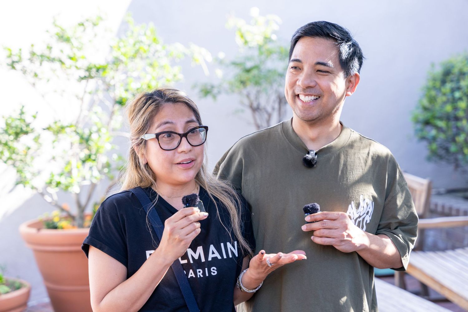 Two people smiling and holding small objects outdoors on a rooftop patio with plants in the background.