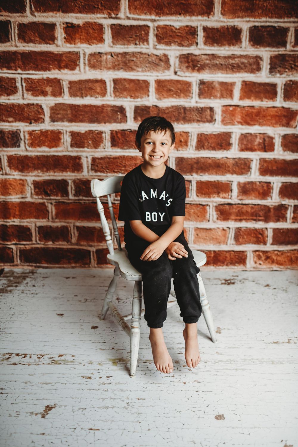 Smiling boy in black Mama's Boy t-shirt sits barefoot on white chair against red brick wall.