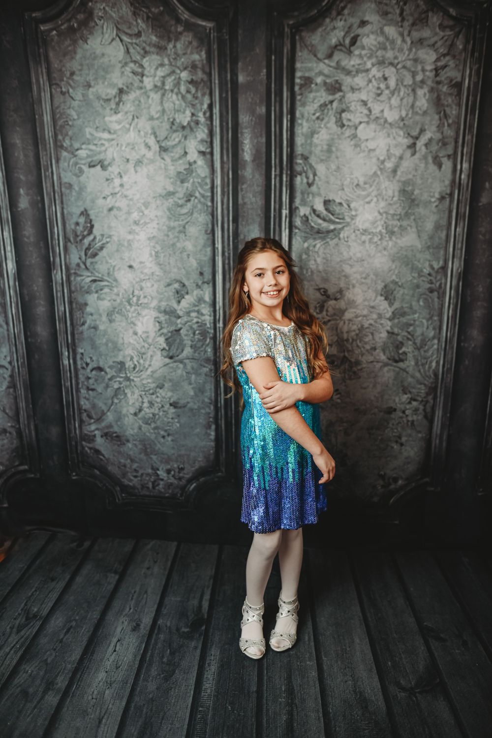 A smiling young girl in a sparkling teal sequin dress poses in front of a vintage floral backdrop.