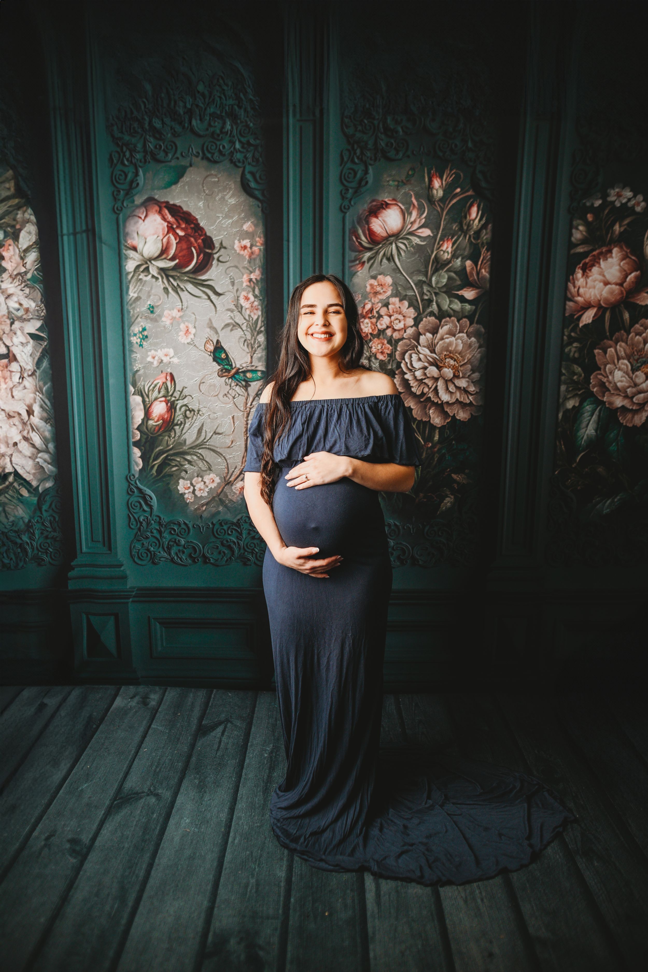 Smiling pregnant woman in navy off-shoulder gown poses against ornate teal floral wall panels on wooden floor.
