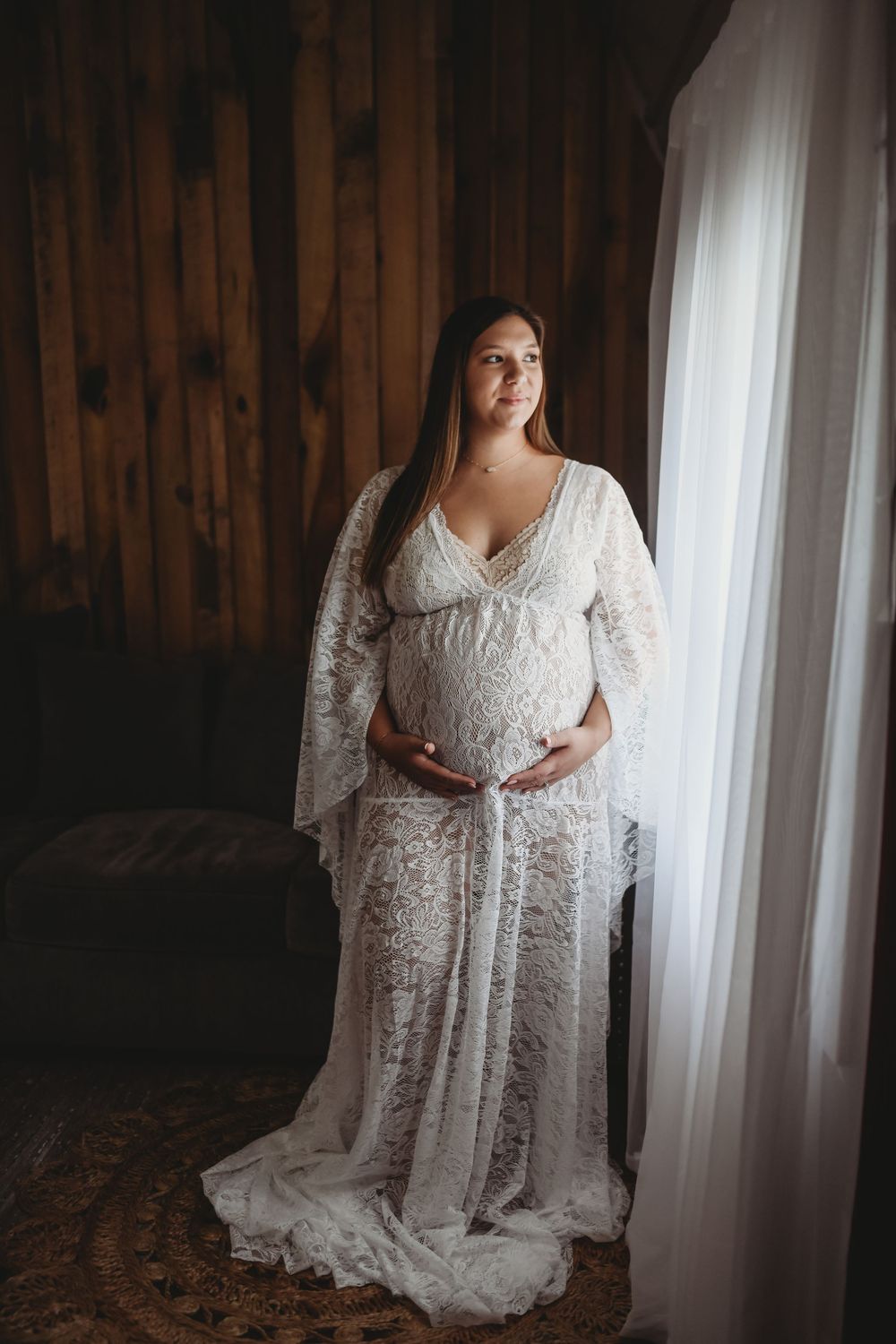 Pregnant woman in white lace maternity gown standing by window in rustic wood-paneled room, cradling baby bump.
