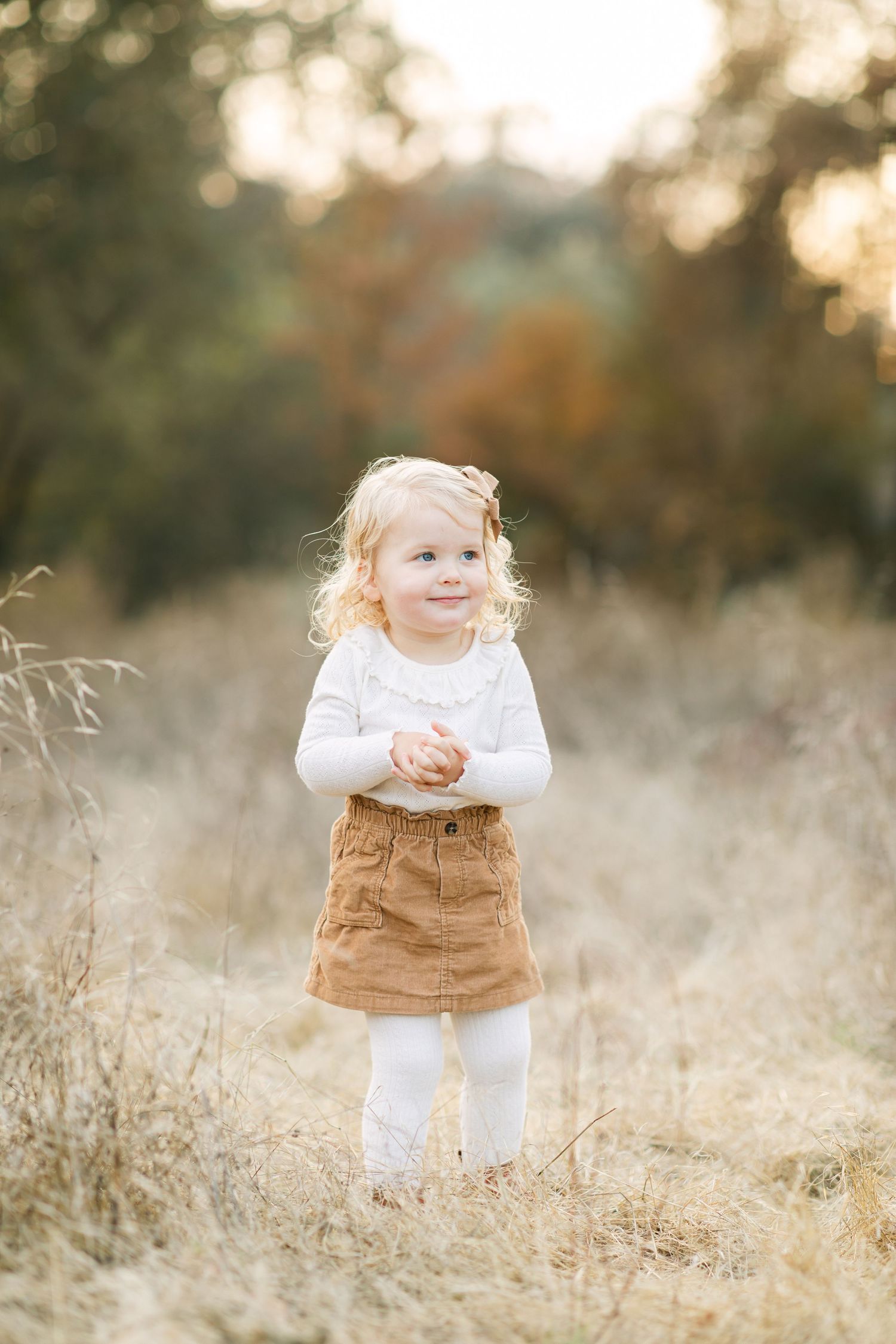 Toddler girl with curly blonde hair wearing a white top and brown corduroy skirt standing in a golden field.