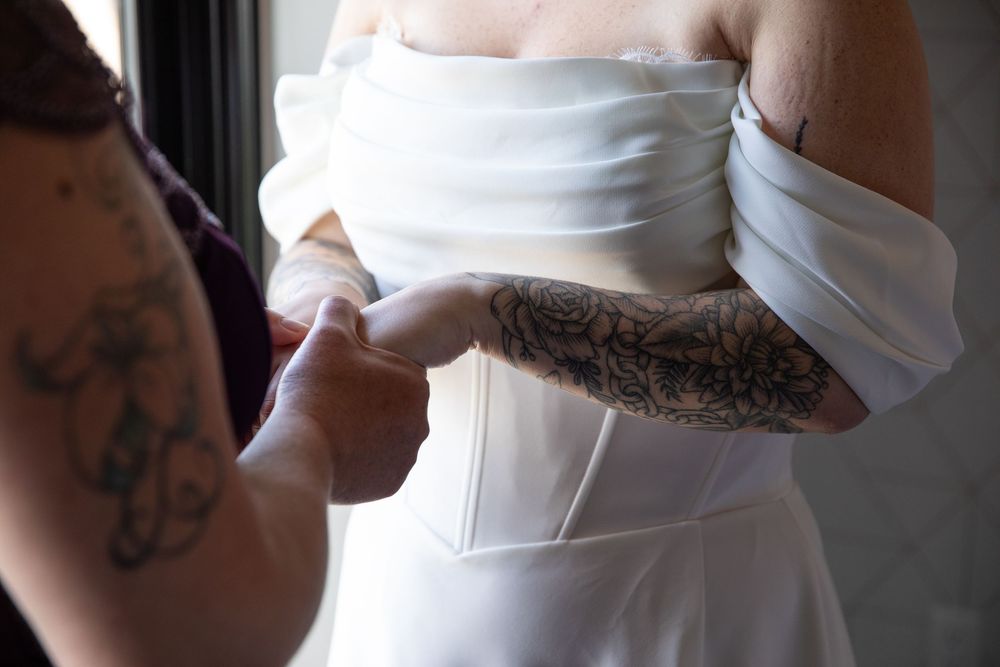 A tattooed bridesmaid helps lace up a bride's white strapless wedding dress before the ceremony.
