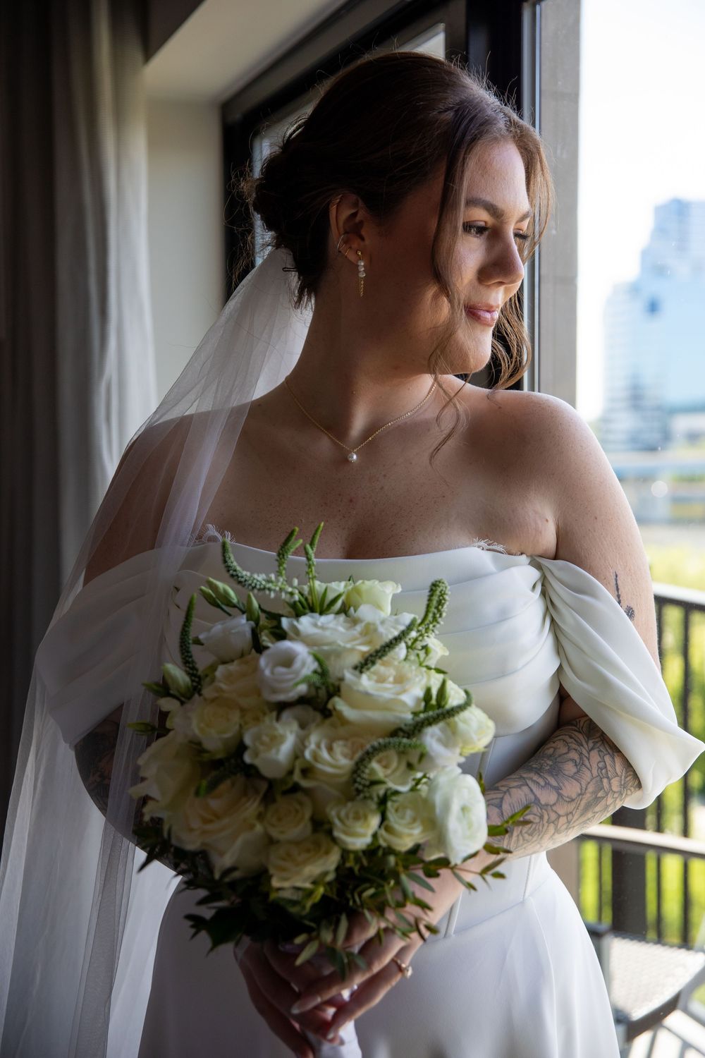 Bride in off-shoulder white gown holding white rose bouquet, wearing veil and tattoos, gazing out window on wedding day.